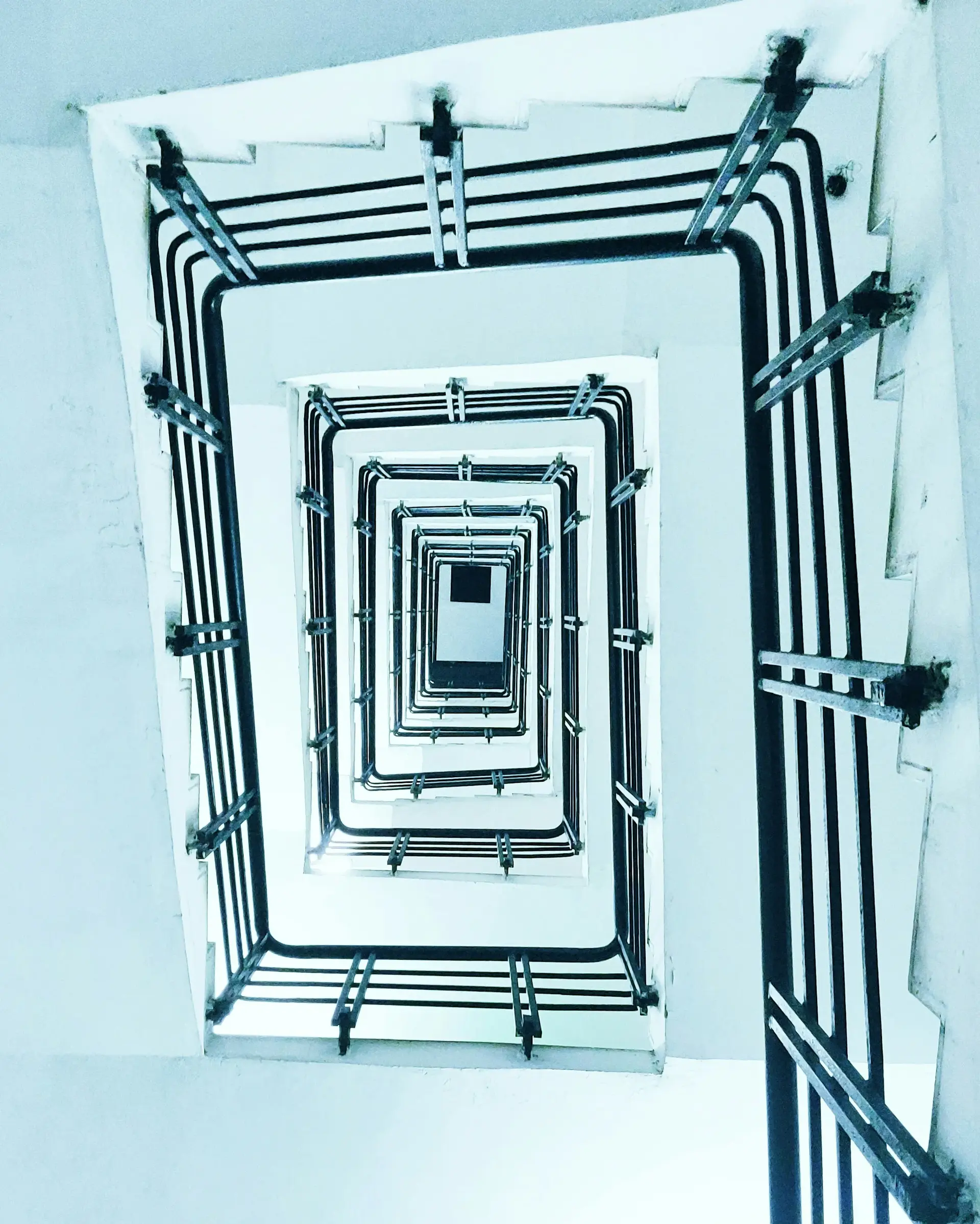 Blick nach oben durch ein quadratisches Spiraltreppenhaus mit weißen Wänden und schwarzen Geländern.