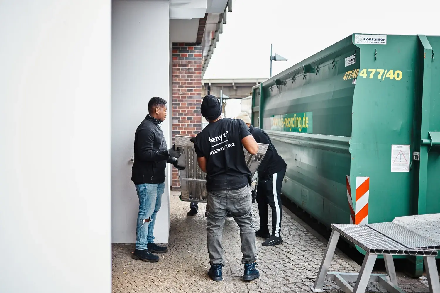 Drei Männer, die im Freien neben einem großen grünen Recyclingcontainer ein Metallobjekt anheben.