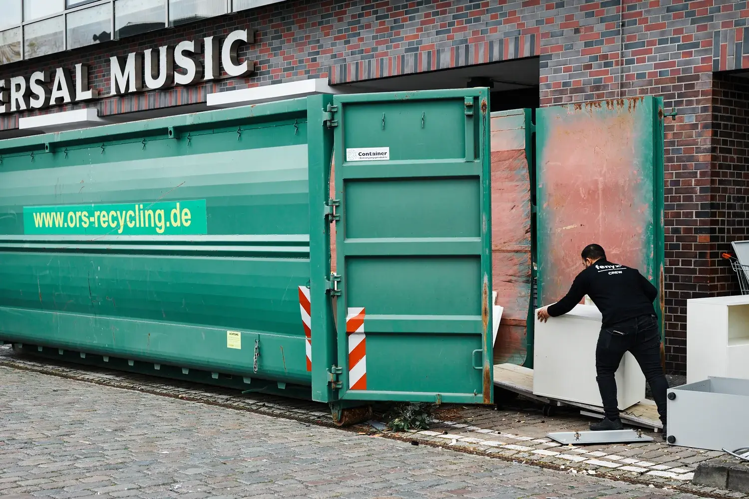Person, die weiße Möbel in einen großen grünen Recyclingcontainer vor einem Backsteingebäude lädt.