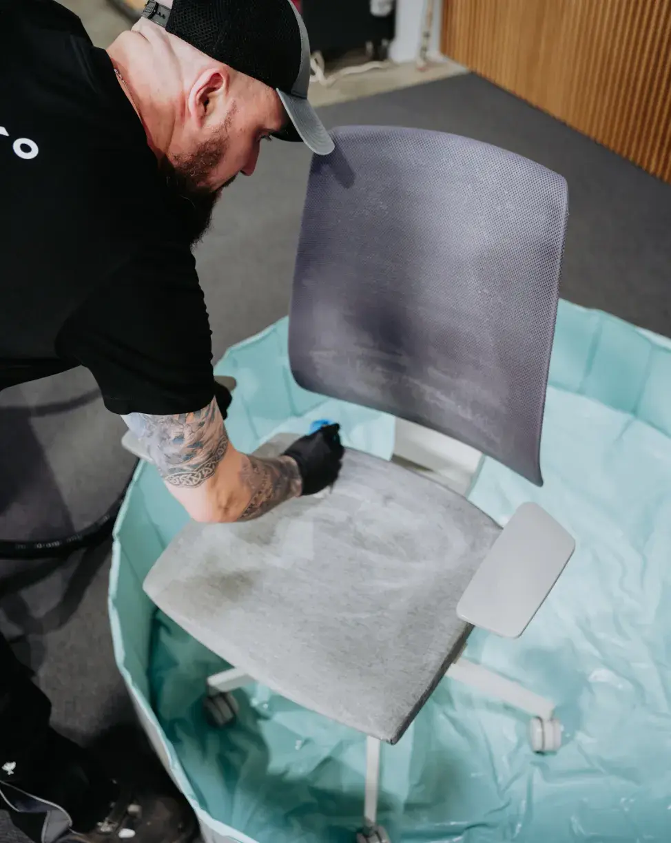 Mann mit schwarzer Mütze und Handschuhen, der einen grauen Bürostuhl in einer blauen Plastikwanne reinigt.
