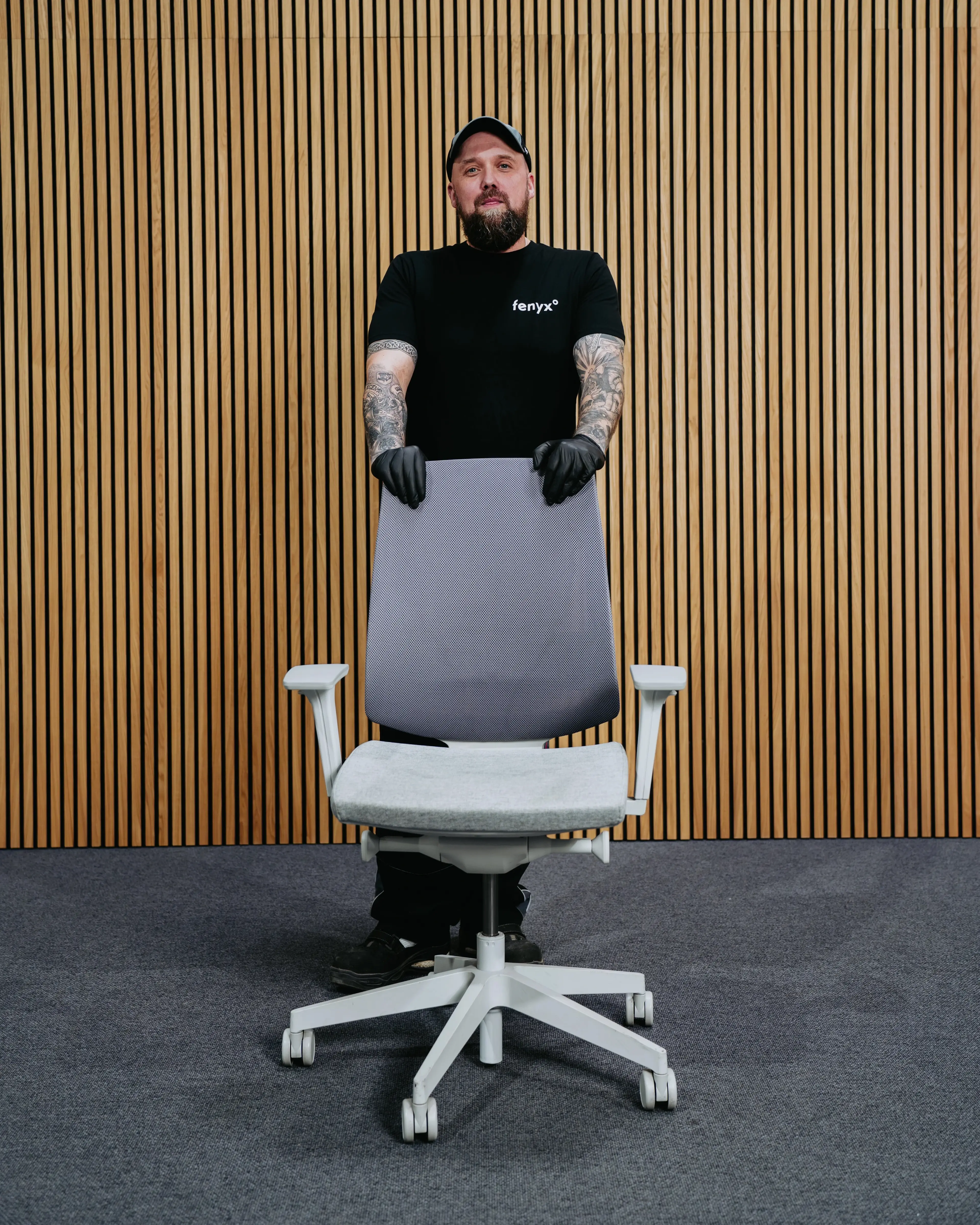 Homme tatoué en gants et t-shirt noirs debout derrière un fauteuil de bureau gris devant un mur à panneaux de bois.