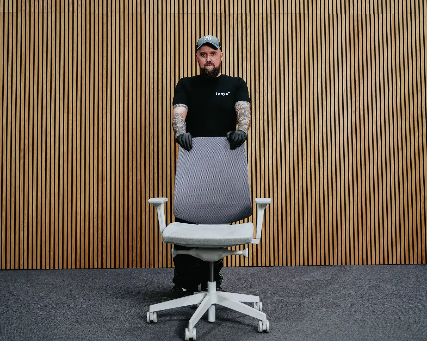 Mann mit Tattoos und schwarzen Handschuhen, der hinter einem Bürostuhl vor einem Holzlatten-Wandhintergrund steht.