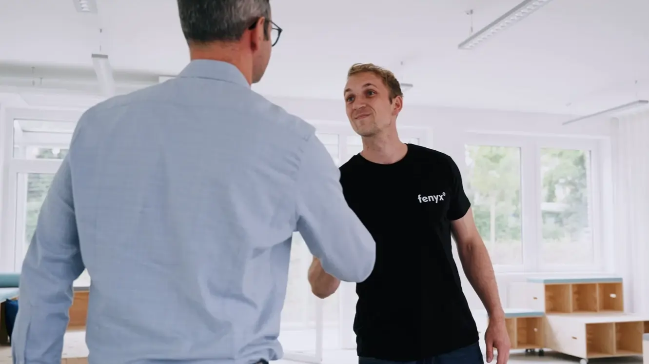 Zwei Männer geben sich die Hand in einem hellen Büroraum, einer trägt ein schwarzes Fenyx-Shirt und der andere ein blaues Shirt mit Brille.