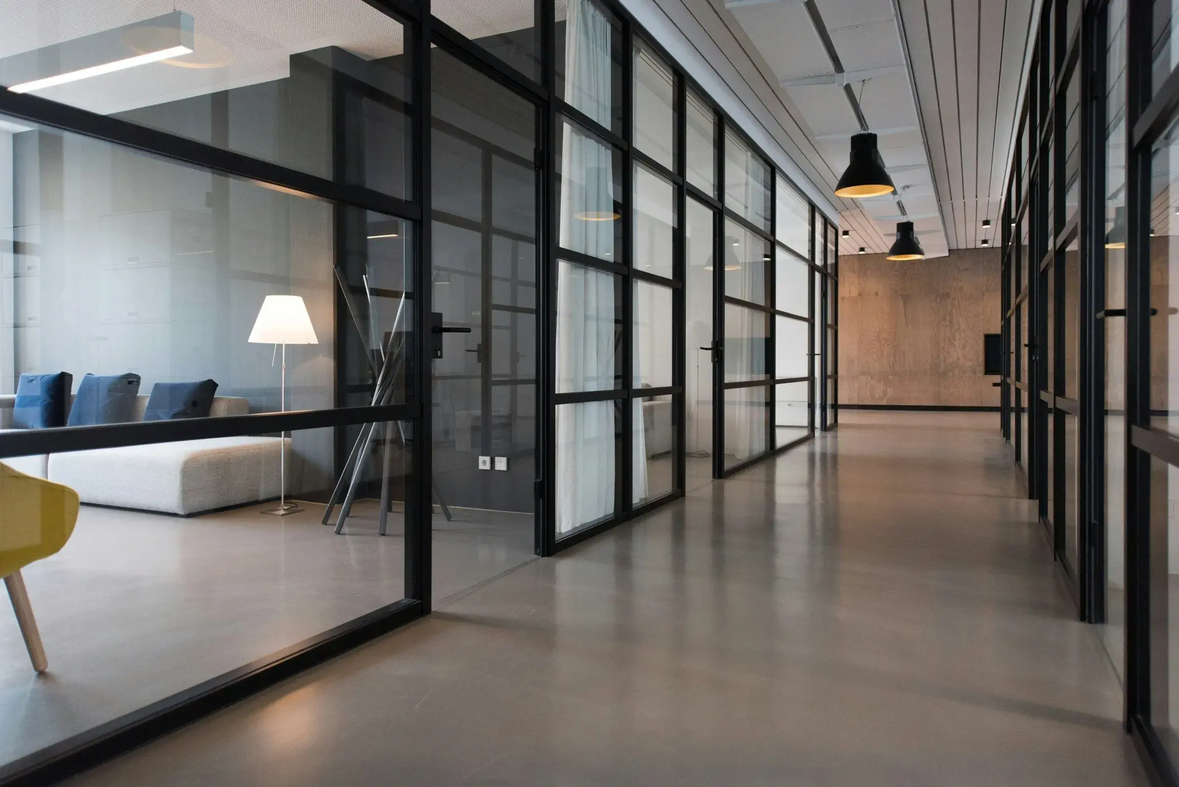Moderner Büroflur mit schwarzen Stahlrahmen-Glaswänden, Betonboden und einem Loungebereich mit gelbem Stuhl im Hintergrund.