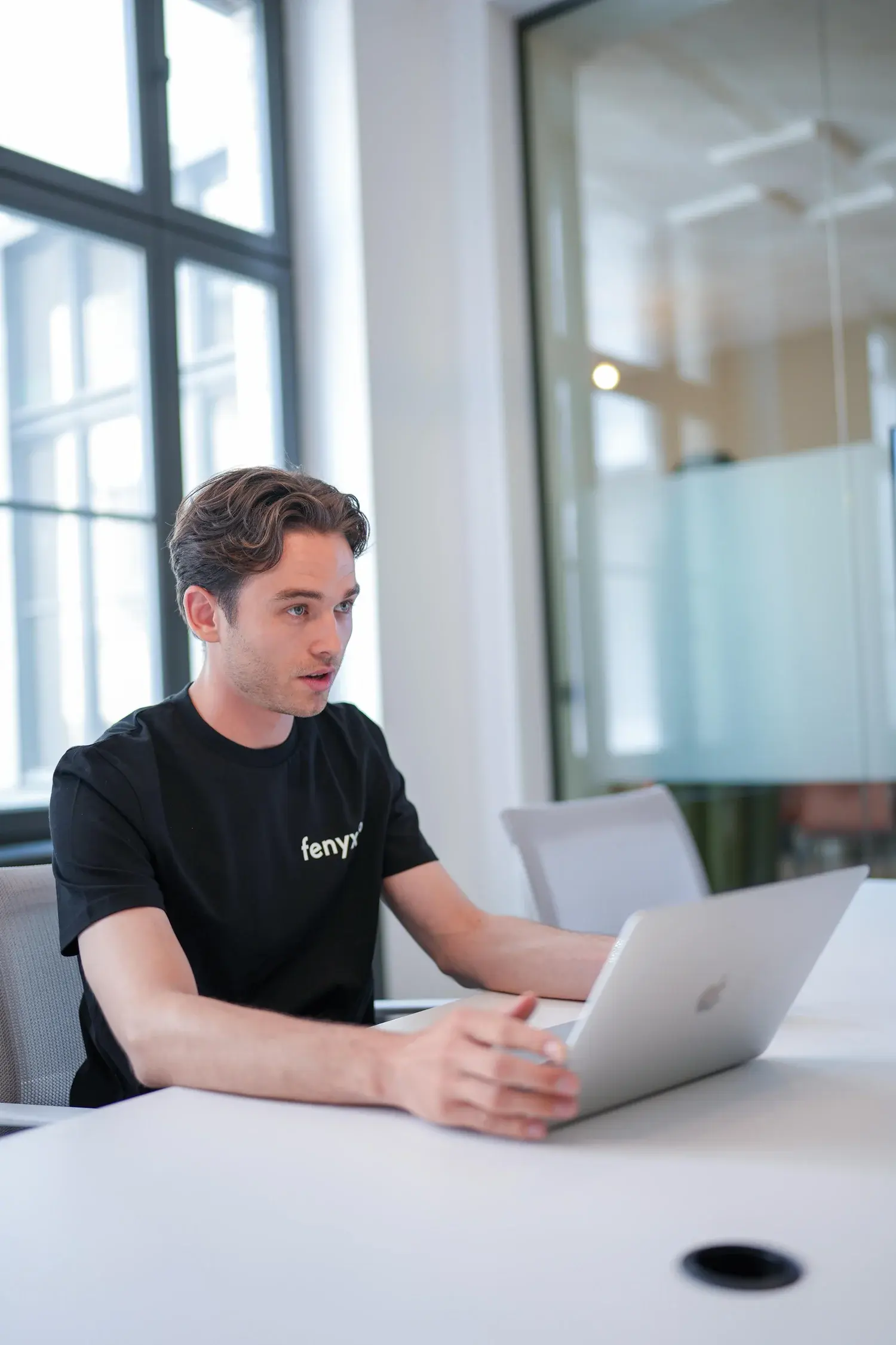 Junger Mann in einem schwarzen Fenyx-T-Shirt, der an einem weißen Bürotisch an einem silbernen Laptop arbeitet.