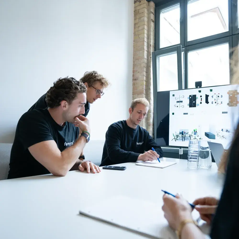 Trois hommes collaborant autour d'une table dans un bureau lumineux, l'un écrivant sur un bloc-notes avec un écran affichant des graphiques en arrière-plan.