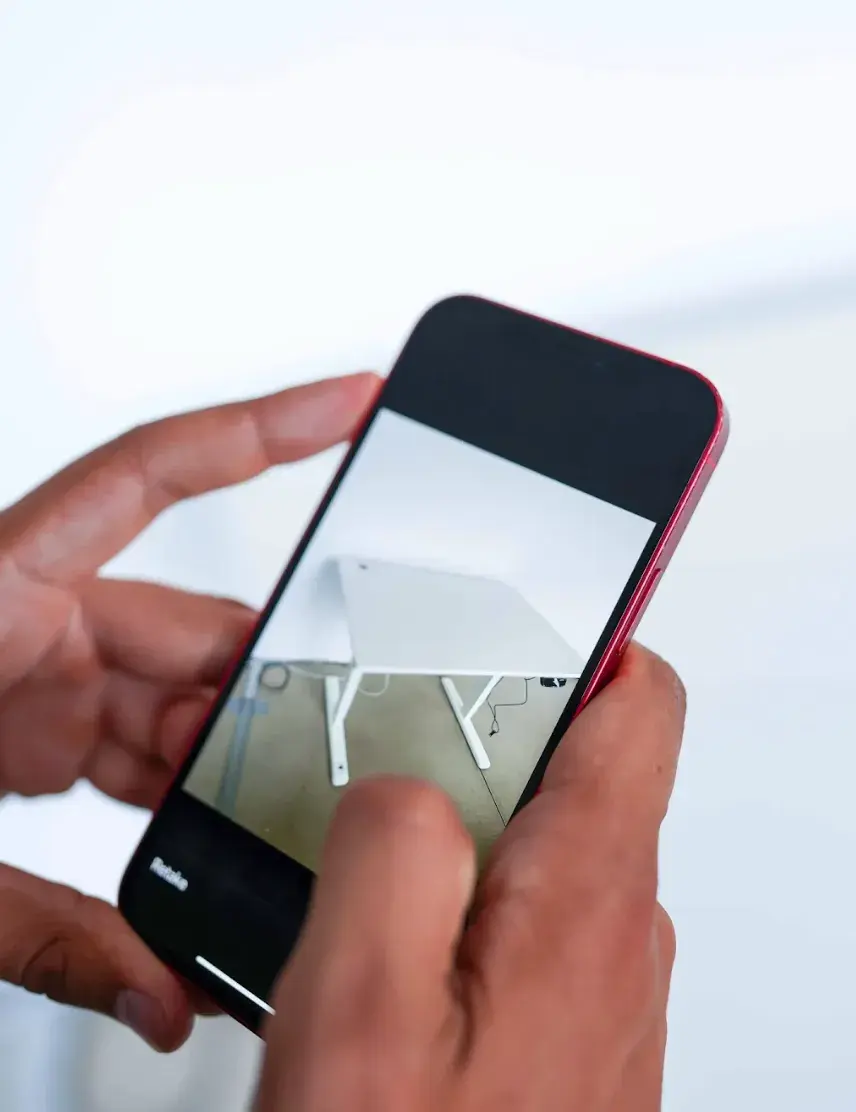 Person, die ein Smartphone hält, auf dem ein Foto eines weißen Schreibtisches mit angewinkelten Beinen vor einer weißen Wand zu sehen ist.