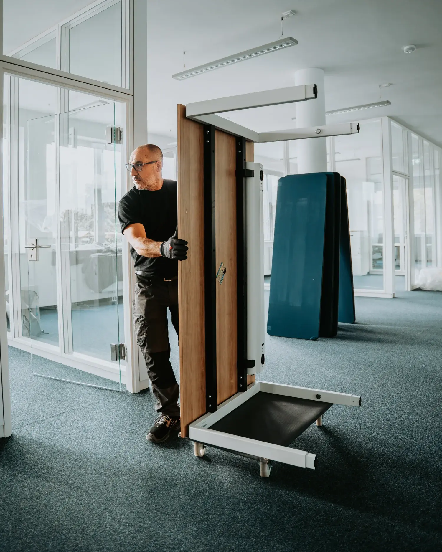Mann mit Brille und Handschuhen, der einen zusammengeklappten Holztisch auf Rollen in einem modernen Büro mit Glaswänden bewegt.