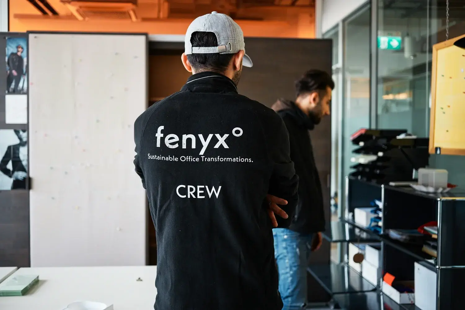 Mann in einer schwarzen Jacke mit dem Aufdruck „fenyx Sustainable Office Transformations CREW" auf dem Rücken, der in einer Büroumgebung steht.