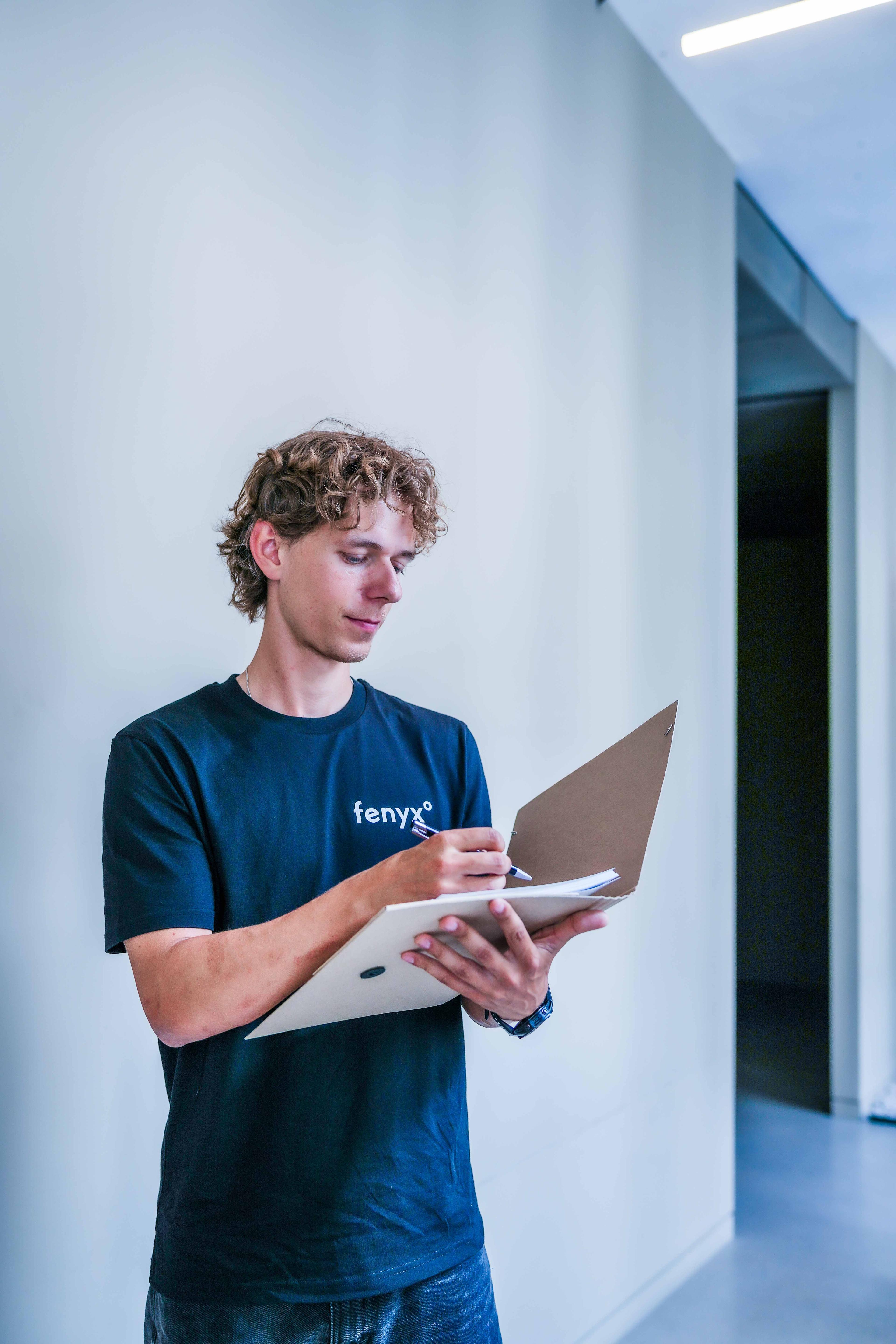 Junger Mann mit lockigem Haar in einem schwarzen Fenyx-T-Shirt, der in einem Flur auf einem Klemmbrett schreibt.